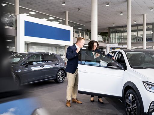 Zwei Personen stehen neben einem Auto in einem Autohaus und haben die Beifahrertür geöffnet. Sie schauen sich den Innenraum des Fahrzeugs an.