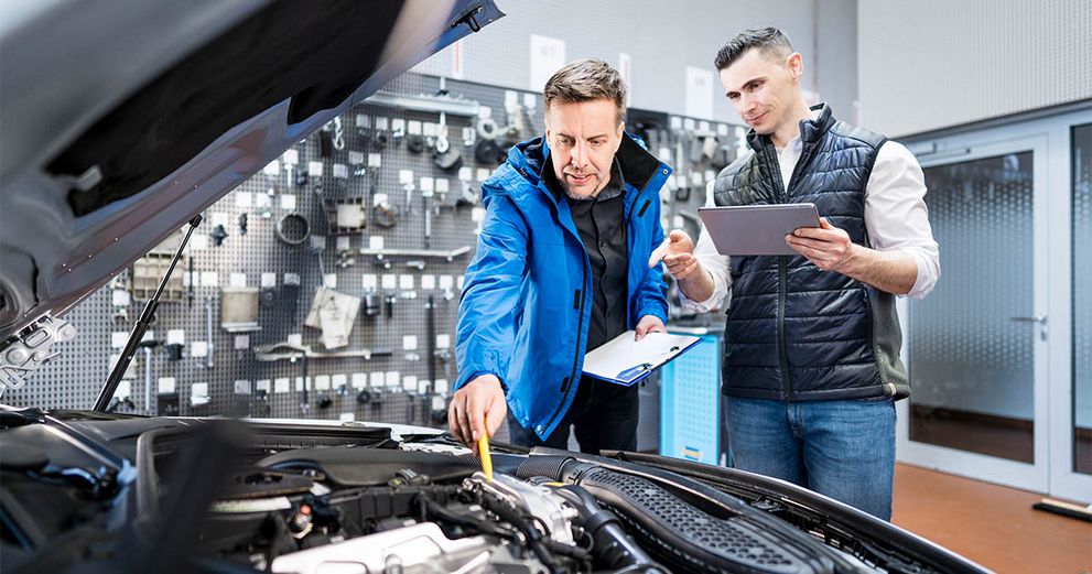 Zwei Personen stehen vor einem Auto mit geöffneter Motorhaube. Eine Person hält ein Klemmbrett in der Hand und zeigt mit einem Stift auf ein Bauteil im Motorraum. Die andere Person hält ein Tablet in der Hand und zeigt ebenfalls mit dem Finger auf das Teil im Motorraum.