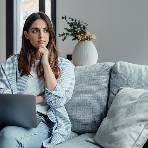 Frau sitzt nachdenklich auf dem Sofa mit einem Laptop in der Hand